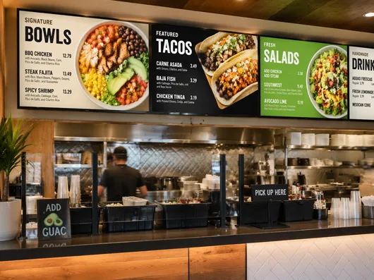 Digital menu boards for bowls, tacos and salads above a restaurant counter